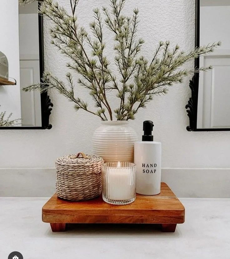“A neatly styled bathroom counter with a ceramic vase holding green branches, a candle, woven basket, and hand soap bottle on a wooden tray, designed with elegance by an Interior Designing Company in Malappuram